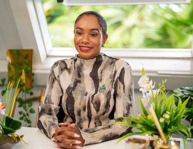 Keithlin Caroo-Afrifa in pattern top sitting at desk surrounded by green indoor plants